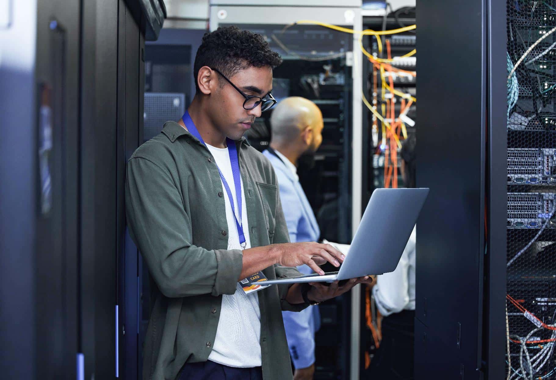 A man working on a laptop in a server room filled with racks and cables