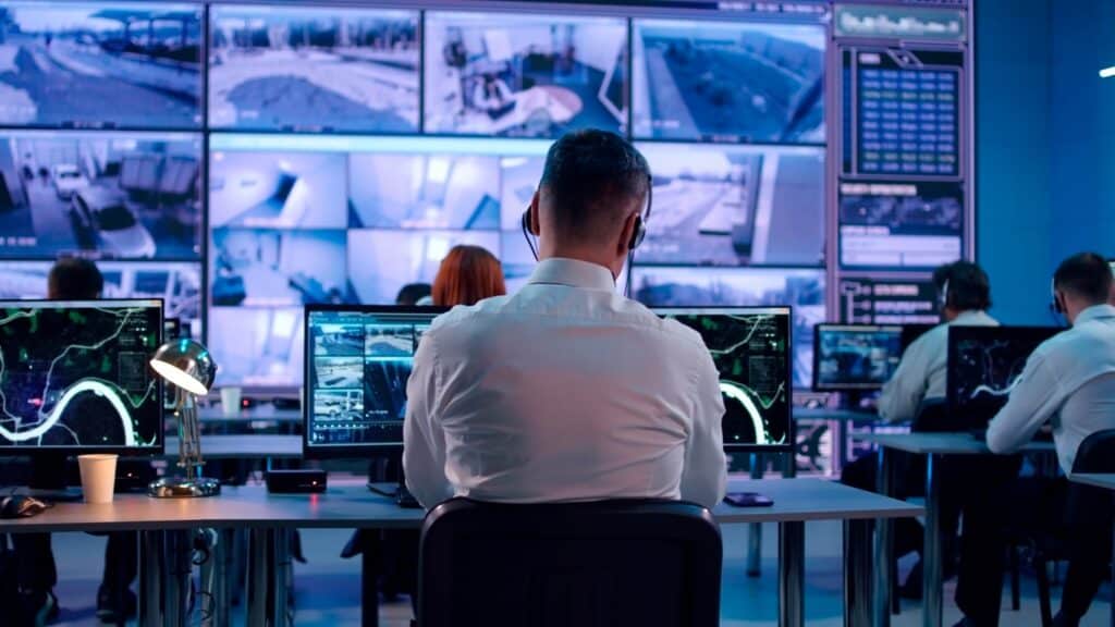 Rows of office workers sit in front of computer monitors which display feeds of security footage. At the front of the room stands a large, wall-sized monitor which displays additional frames of surveillance video.