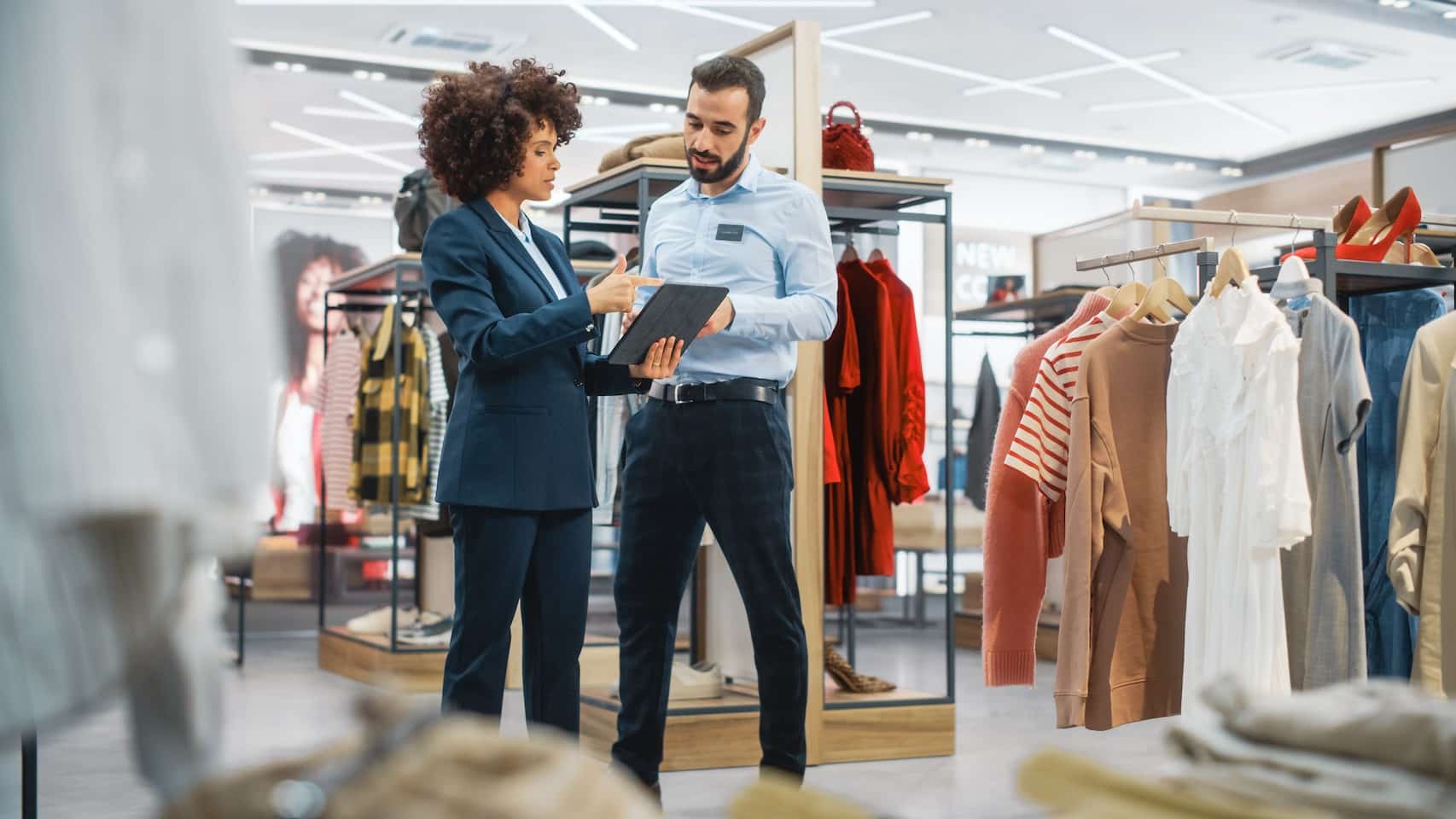 A man and woman in a clothing store look at a tablet held by the woman.