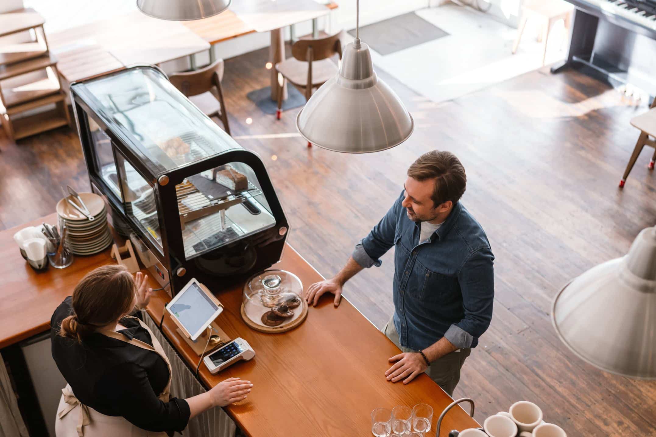 A man stands at a coffee shop counter, engaged in conversation with a female barista.