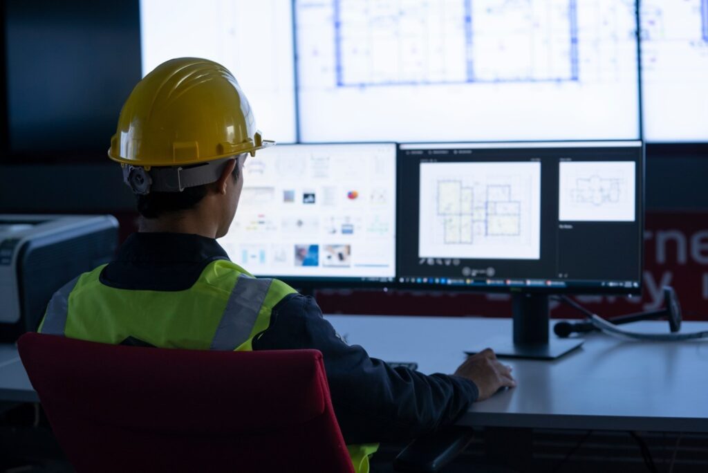 A man wearing a hard hat and safety vest is focused on two computer screens while working on a project.
