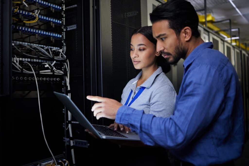 Two people look at a laptop screen while standing in a server room