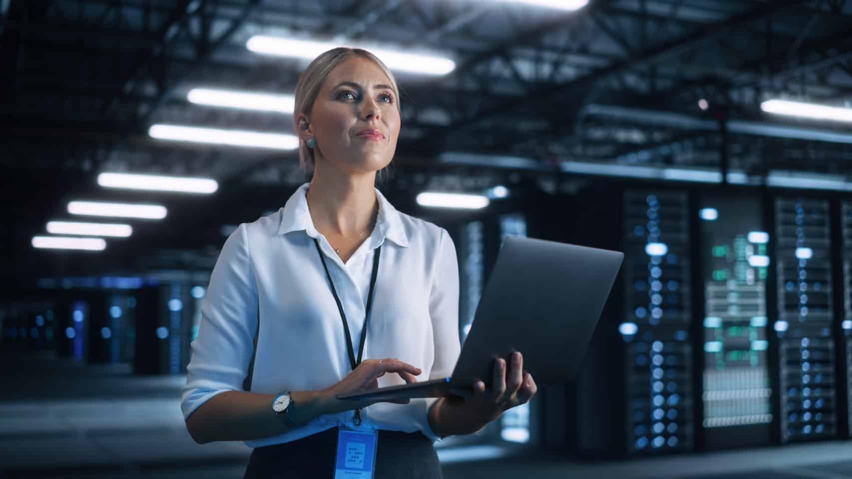 A woman in a business suit holds a laptop while standing in a server room filled with servers and equipment
