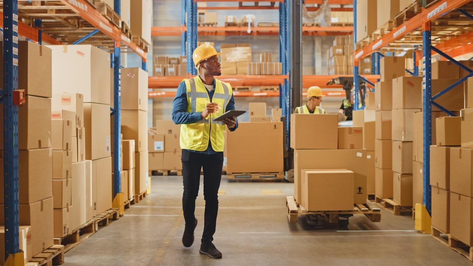 A man wearing a safety vest and hard hat stands in a warehouse, overseeing operations and ensuring safety.