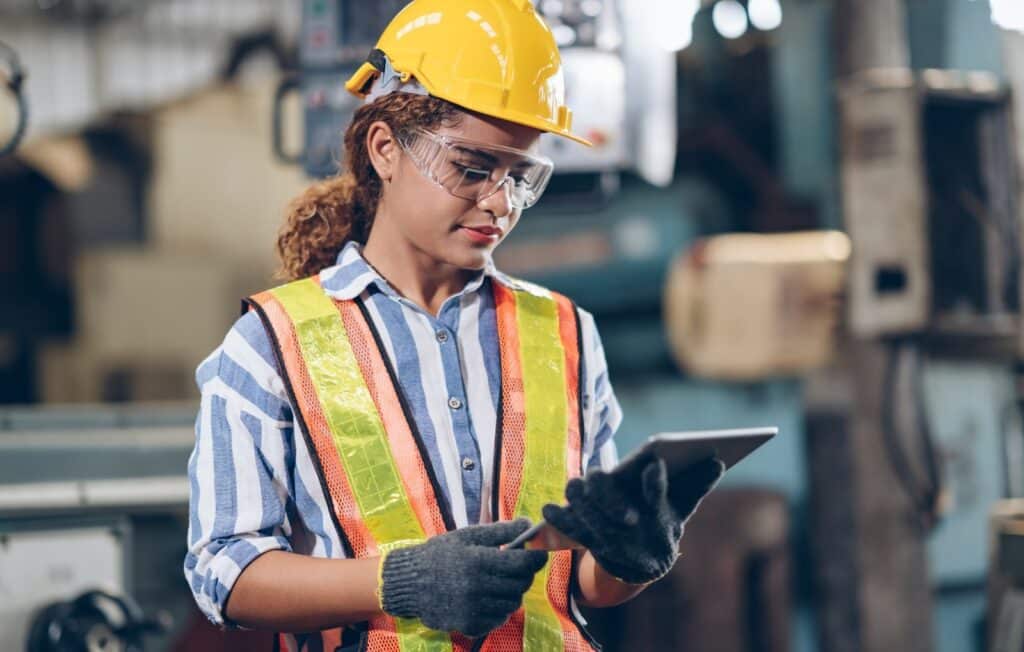 A woman wearing a safety vest and hard hat holding a tablet in a construction or industrial setting.