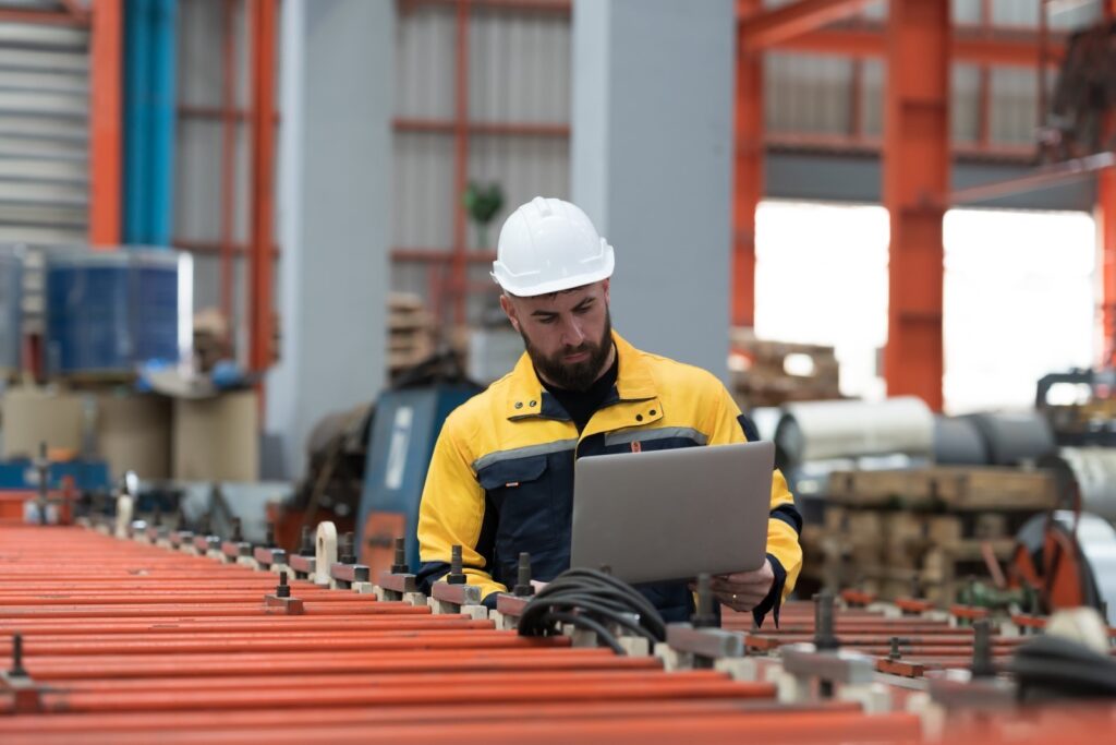 A man in a hard hat works on a laptop while standing on a manufacturing floor