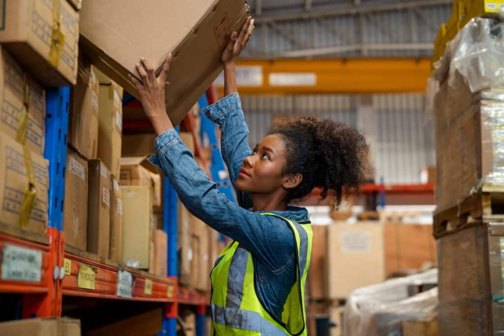 A woman stands in a warehouse, loading a box onto a shelf.