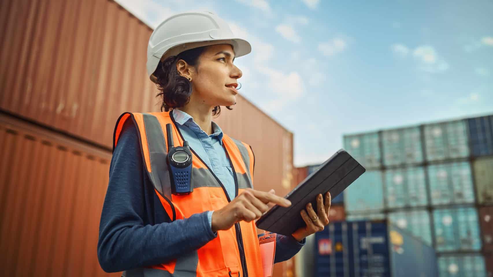 A woman wearing an orange vest and hard hat holding a tablet while standing near shipping containers.