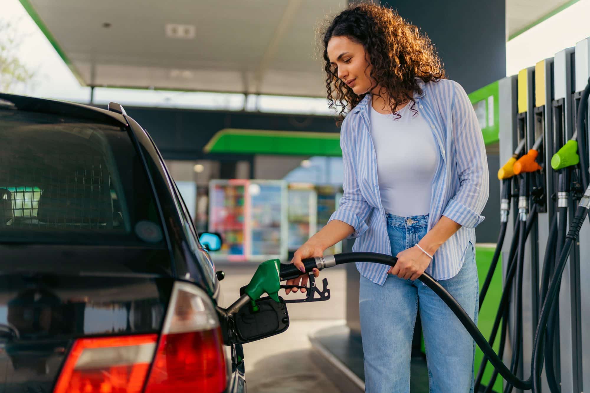 A woman is filling her car with gas at a gas station, holding the fuel nozzle while standing next to her vehicle.