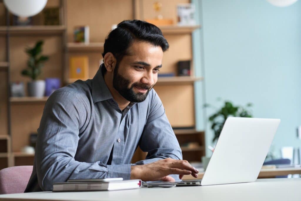 A male teacher sits at his desk, focused on his laptop 