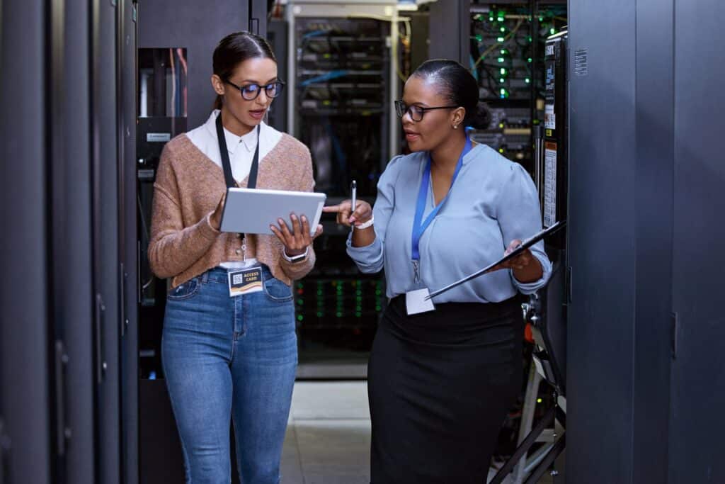Two women stand in a server room examining a tablet, surrounded by servers and networking equipment.