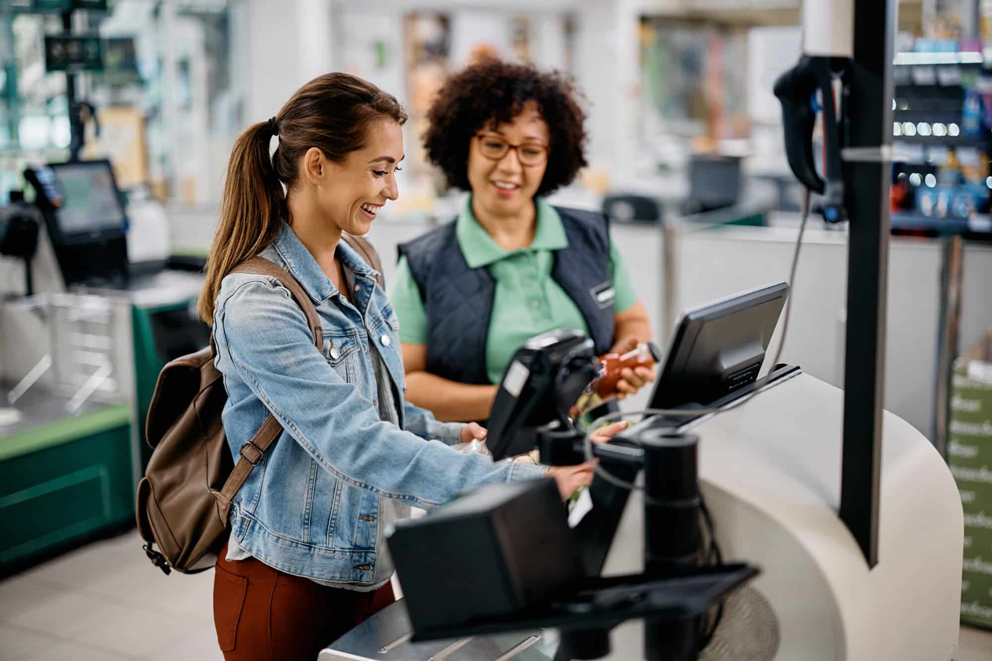 Two women stand at a cash register, one holding items while the other looks on, both smiling.