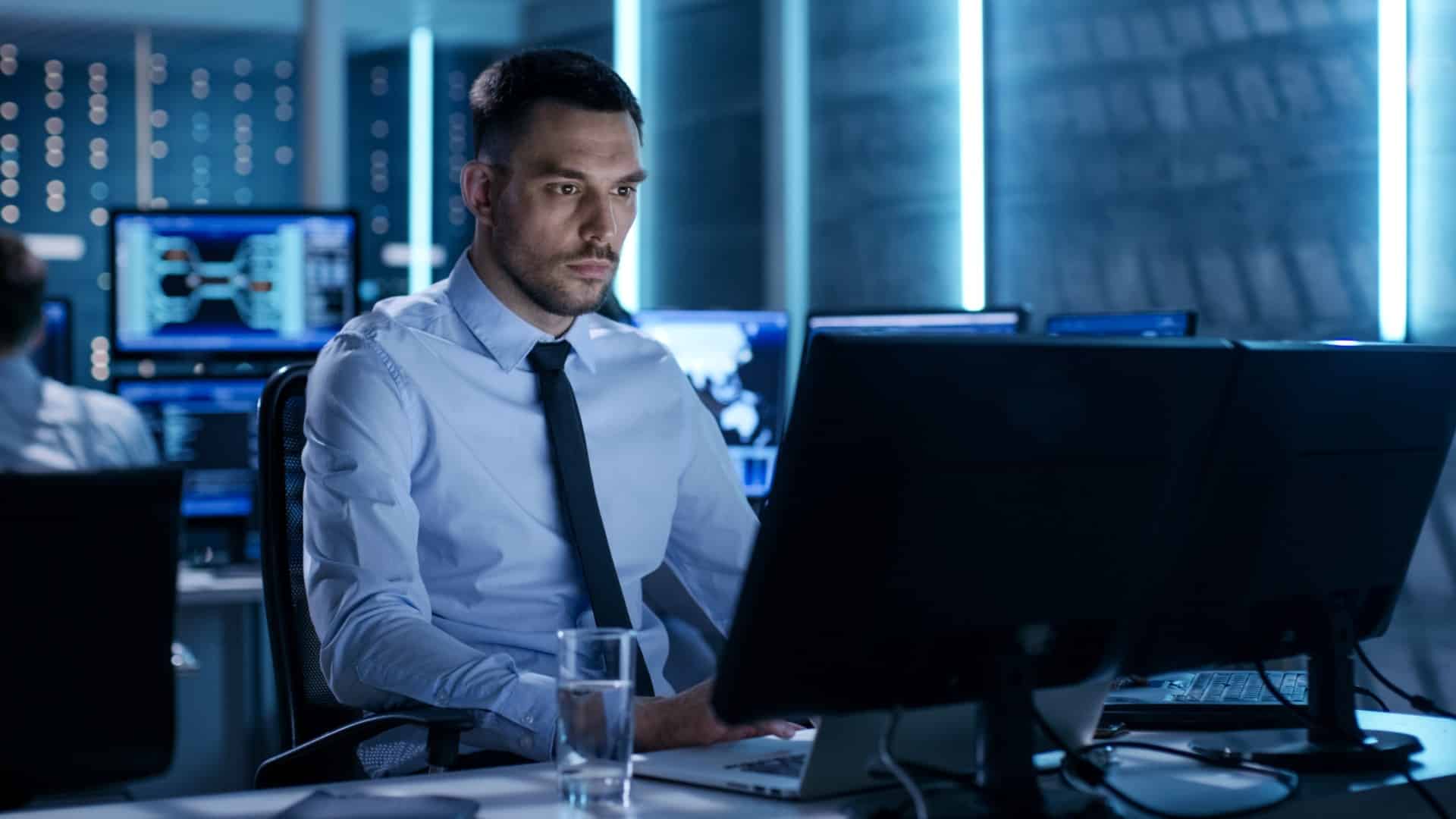A man in a collared shirt and tie sits at a desk, focused on a computer monitor