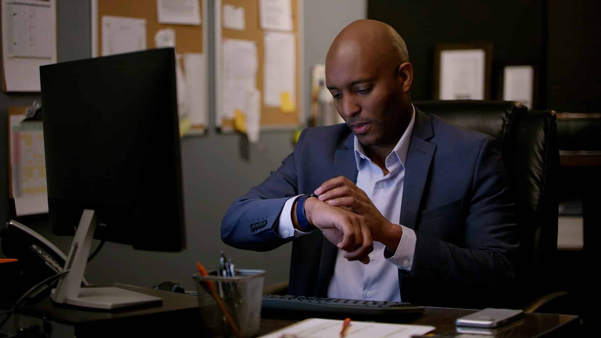 A man in a suit sits at his desk, looking at his watch.