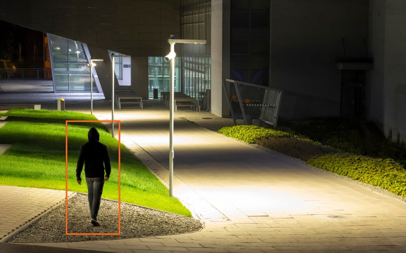 A person in a hoodie walks on a college campus sidewalk at night, highlighted by an orange detection box.