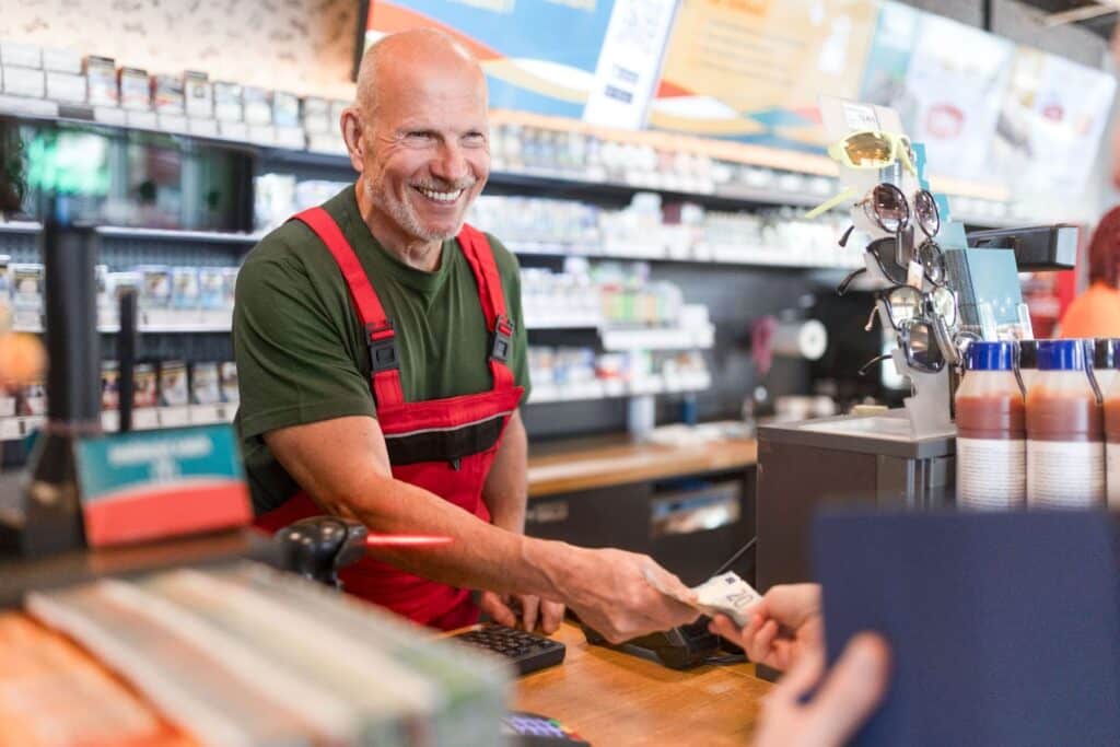 A smiling man stands behind a convenience store counter, handing change back to a customer
