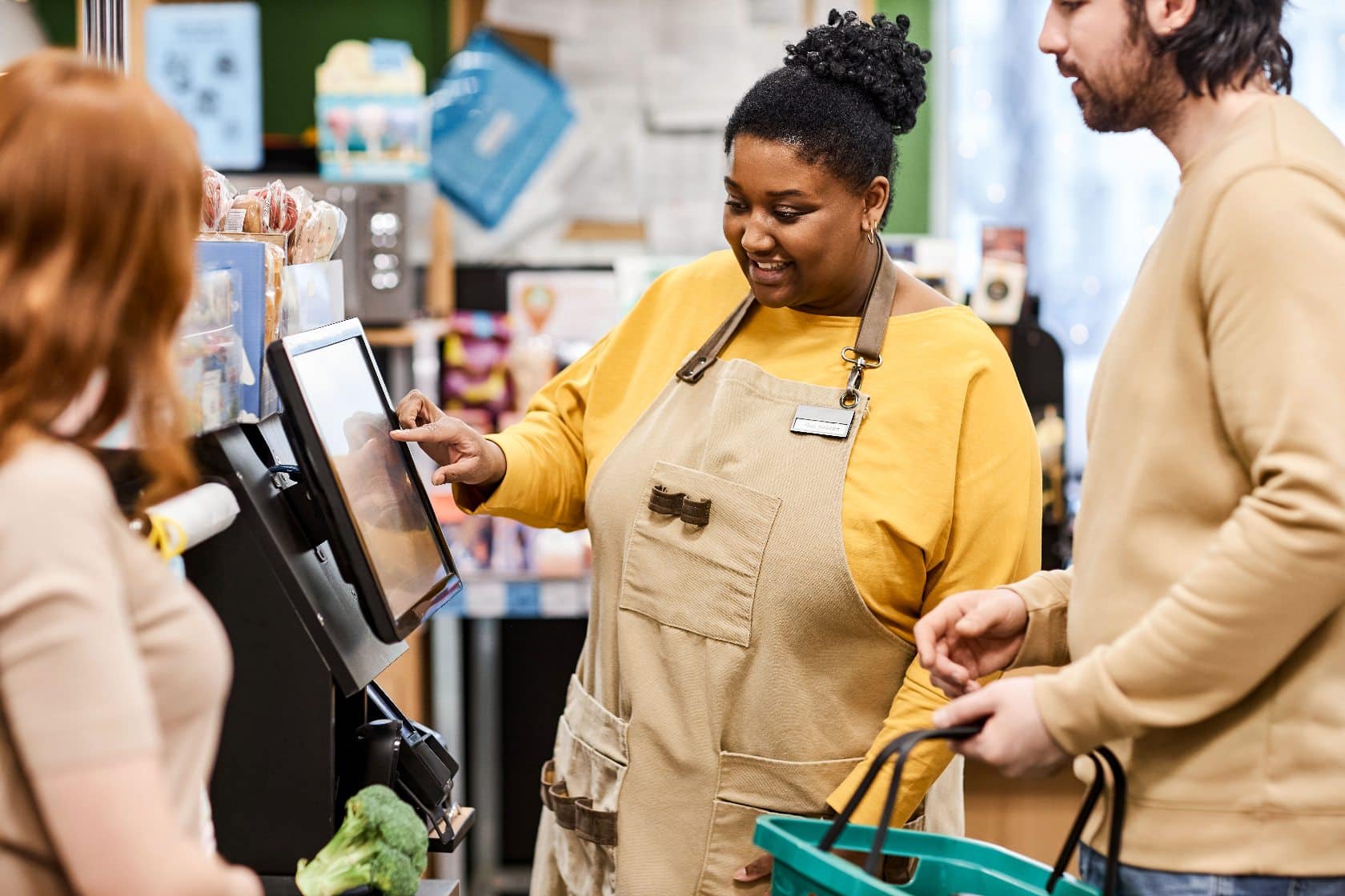 A store employee helps a woman and a man at a self-checkout register in a retail environment.