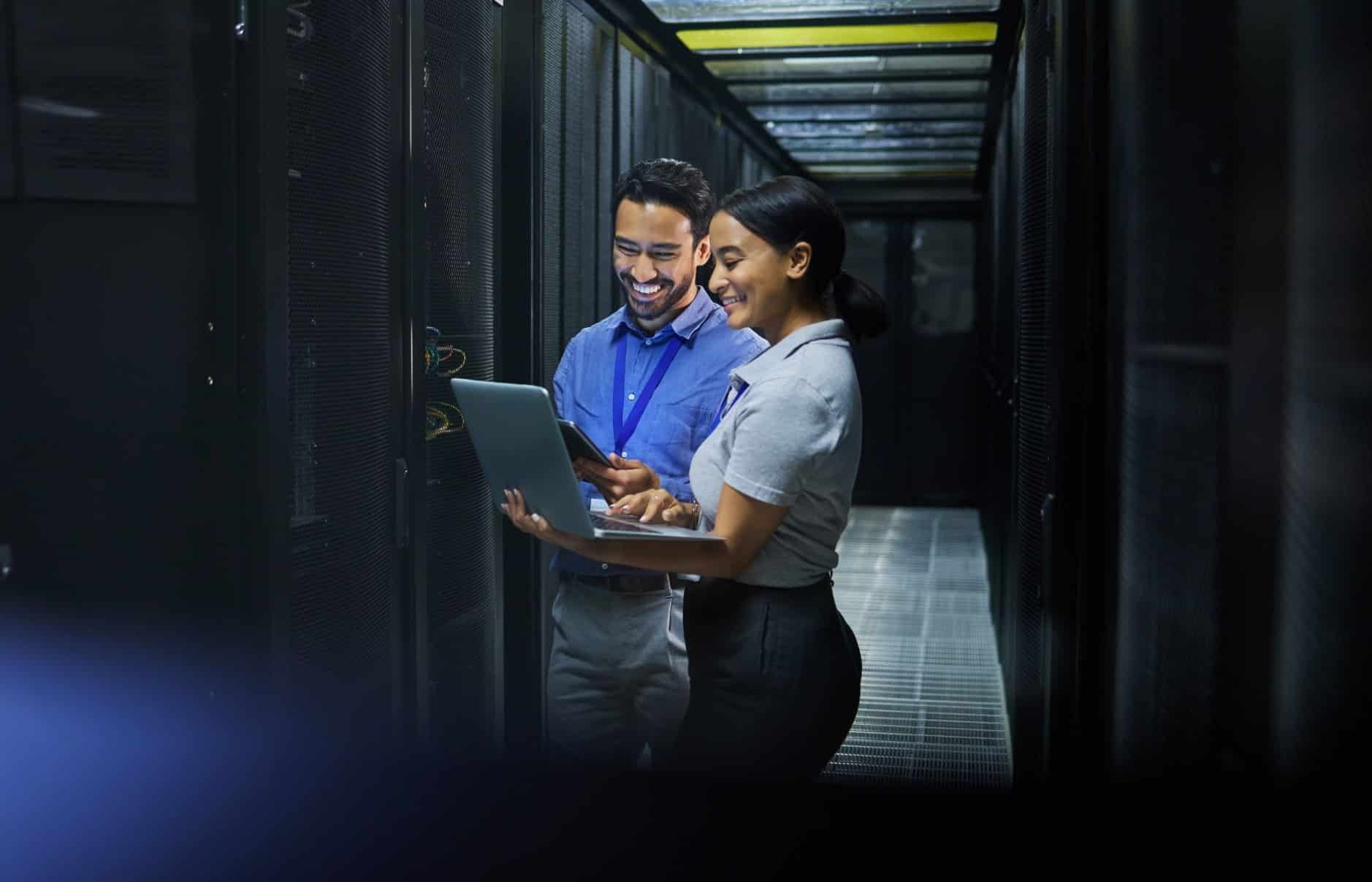 Two individuals in a server room, examining a laptop while surrounded by rows of servers and equipment.