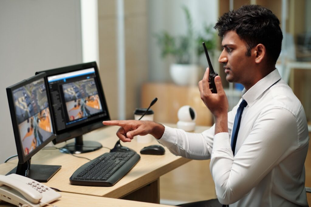 Man sitting at desk with radio communicating what he's seeing on computer screen, which displays OpenEye Web Services video security feeds.