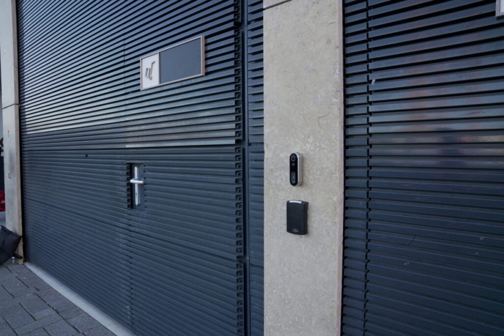 Access control panel and doorbell camera on concrete pillar next to a black door.