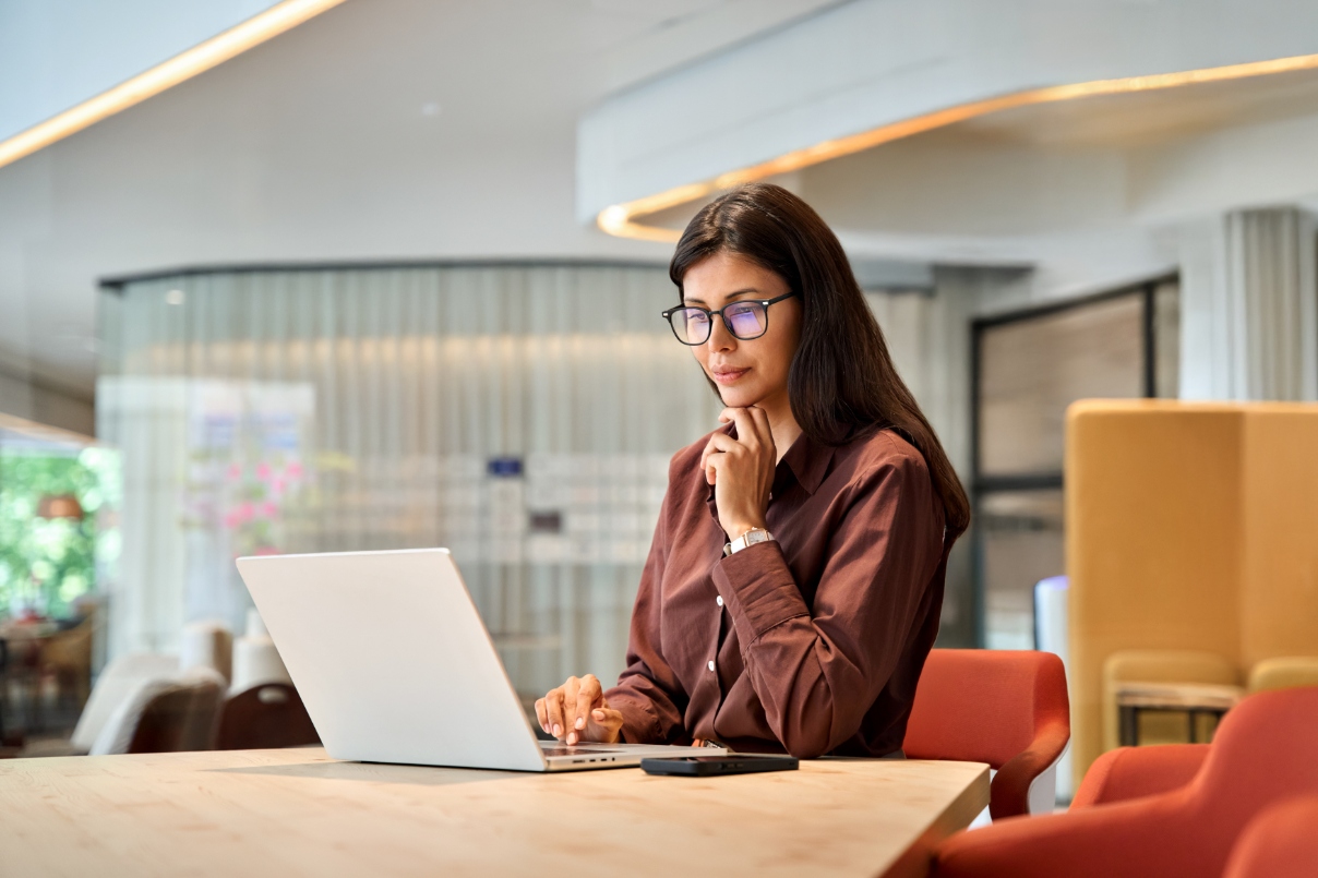 A woman with glasses is seated at a table, engaged with her laptop, indicating a work or study environment.