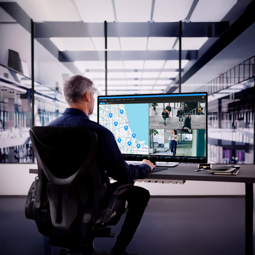 Man in office setting viewing computer displaying live camera feeds and cloud-based maps in OpenEye Web Services.