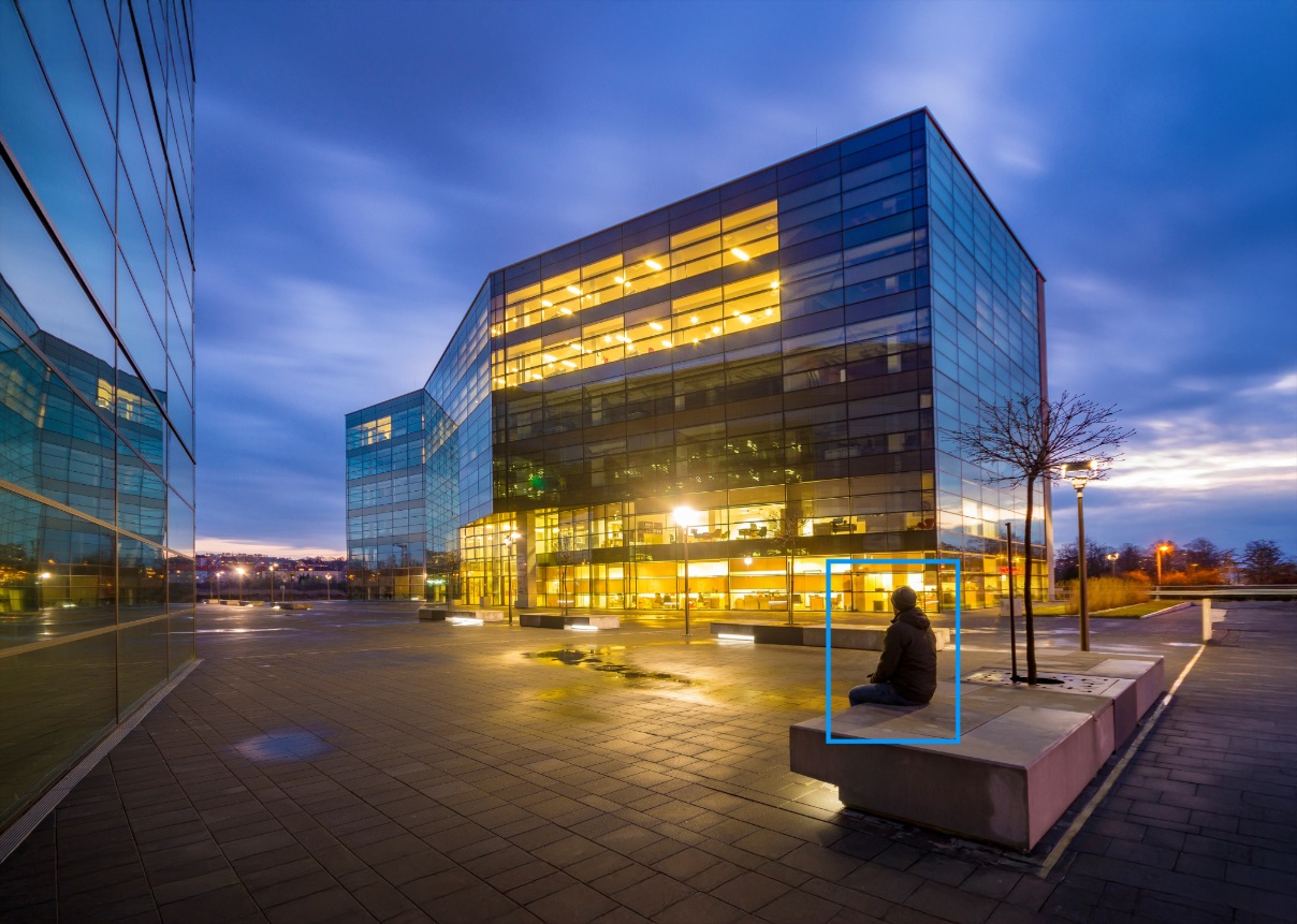 Individual sitting outside of building at night with blue bounding box generated by AI video analytics around them.