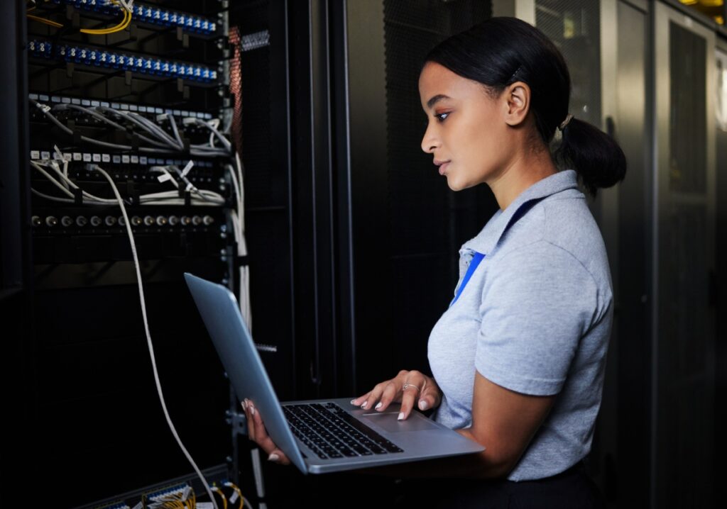 Woman standing next to multiple servers holding a laptop.