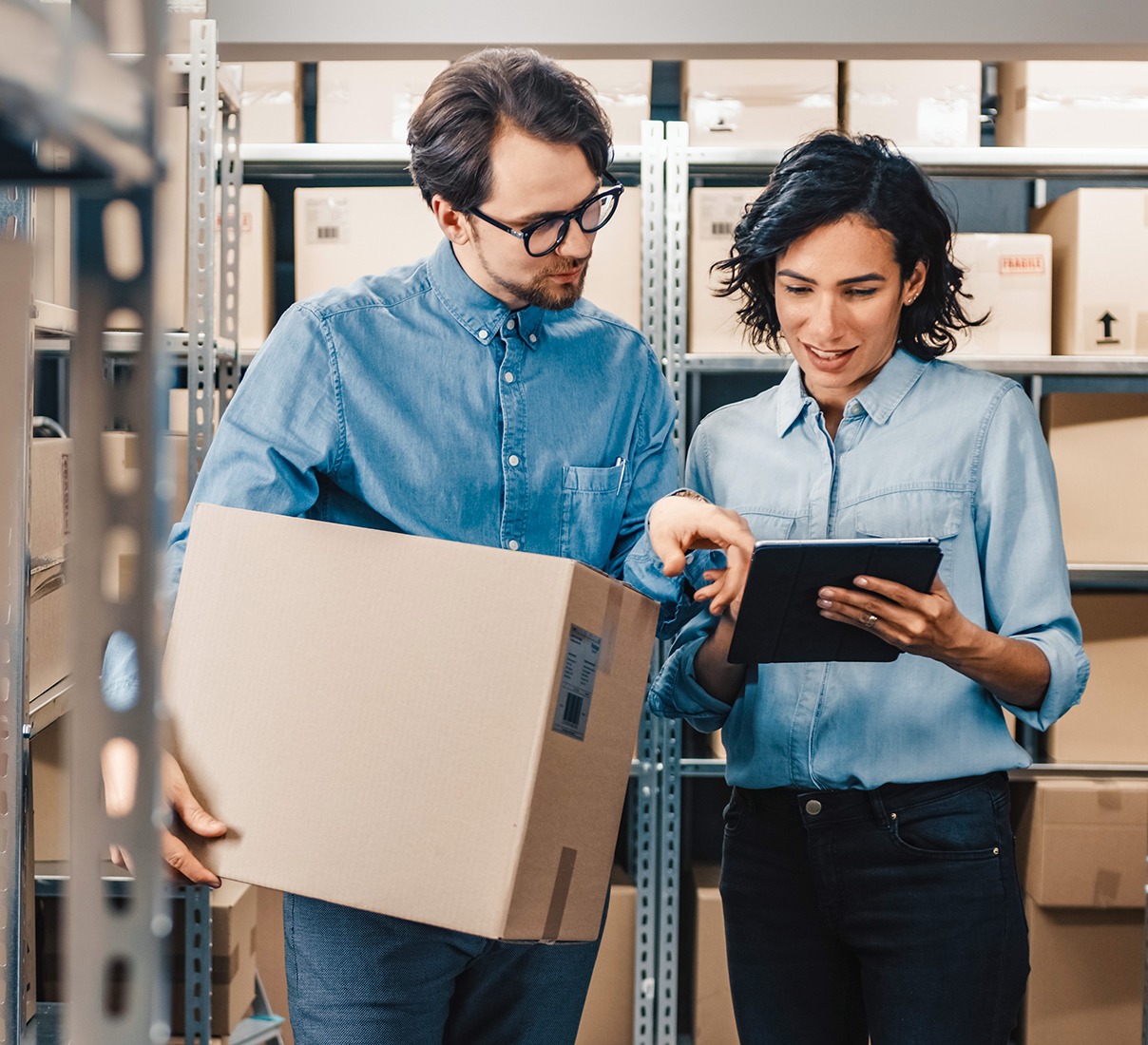 Two individuals stand in a warehouse surrounded by stacked boxes, indicating an active storage environment.