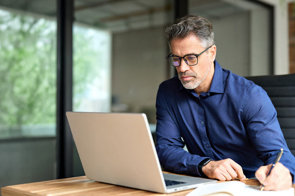 Man sits at desk writing with a pen while he looks at a computer screen.