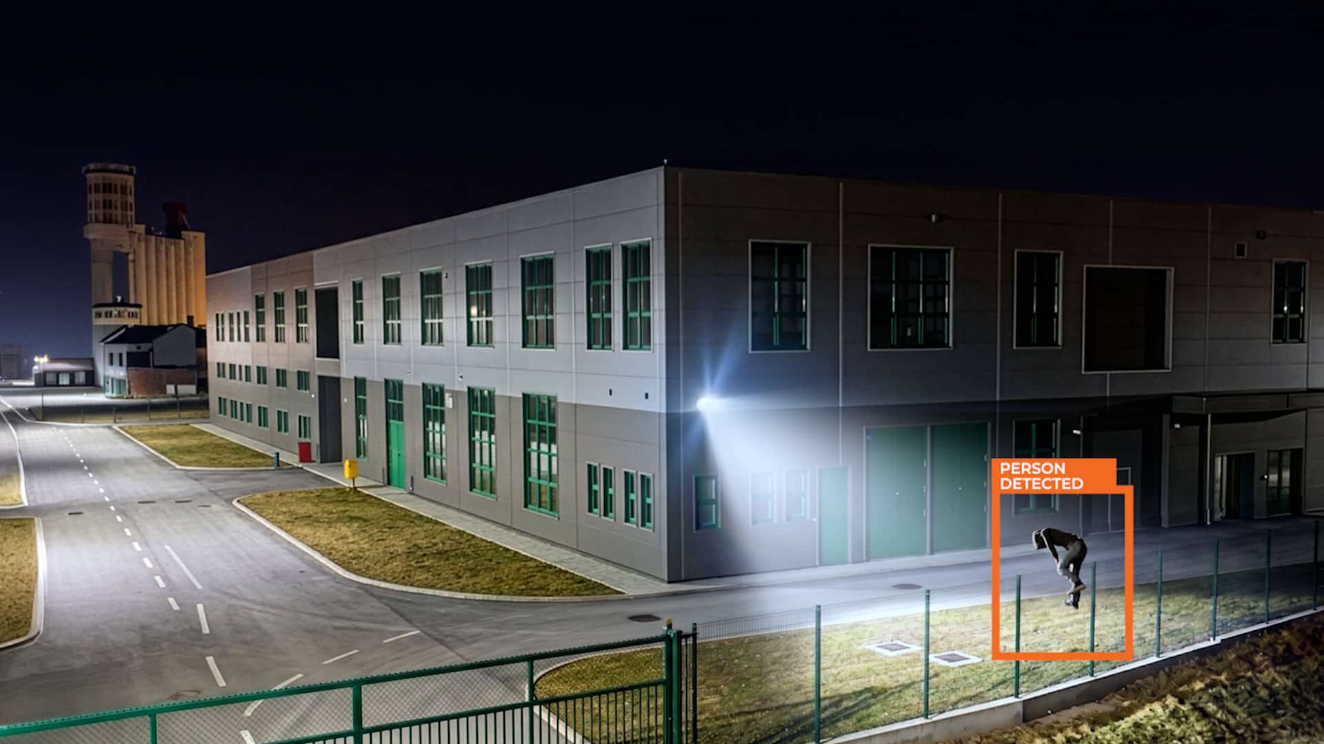 A person attempting to break into a warehouse is illuminated from an OpenEye Active Deterrent camera. The individual has an orange bounding box surrounding them.