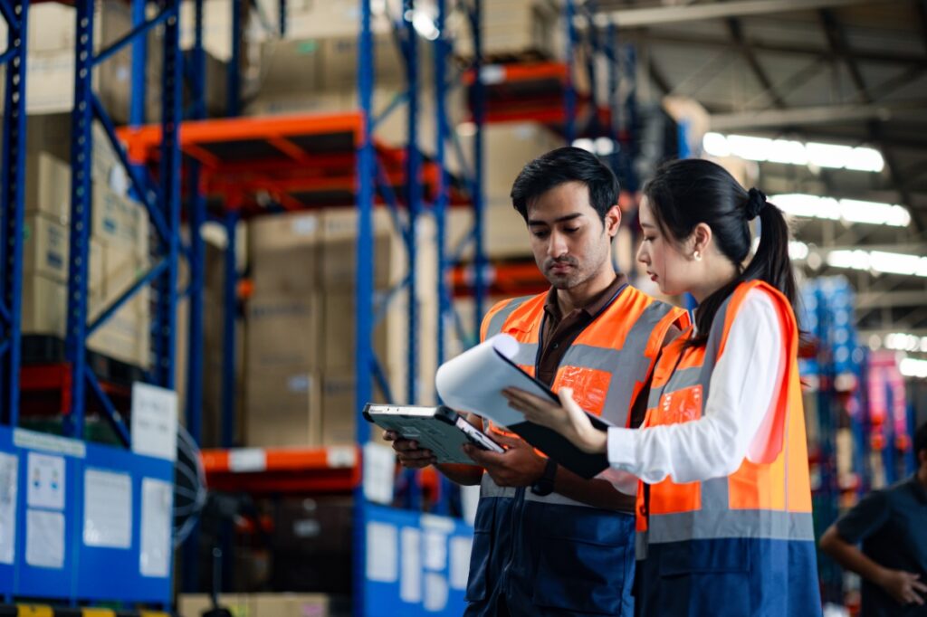 A man holding a tablet and a woman holding a clipboard conduct a manual walkthrough in a warehouse setting.