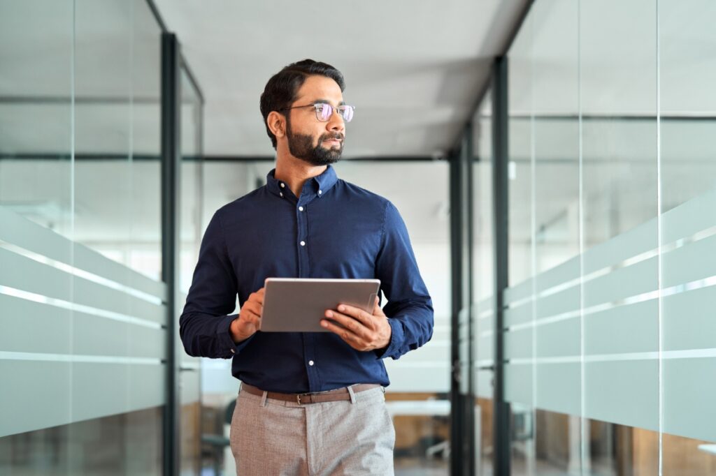 Man stands in office setting looking through a window and holding a tablet in his hands.