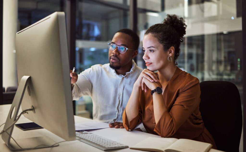 Two people sit and look at a computer screen in an office setting.