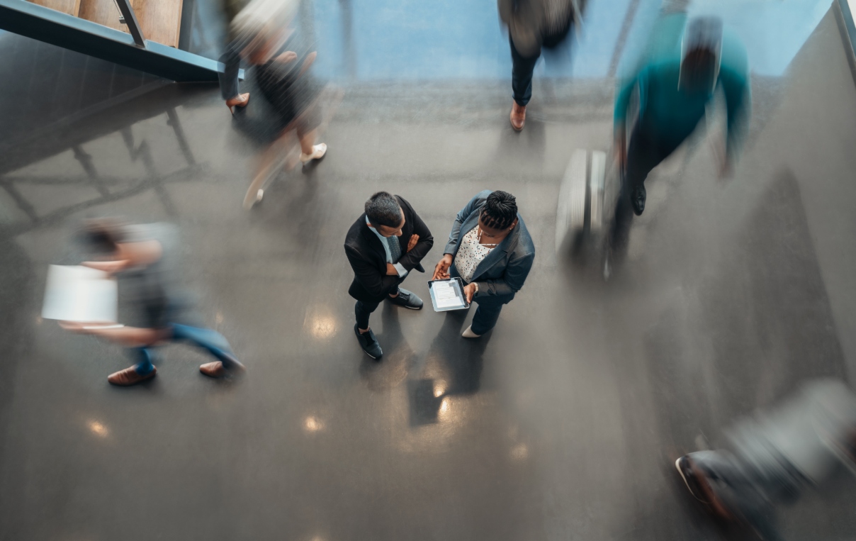 Two individuals stand in an office setting inspecting a tablet while they are surrounded by passing people.