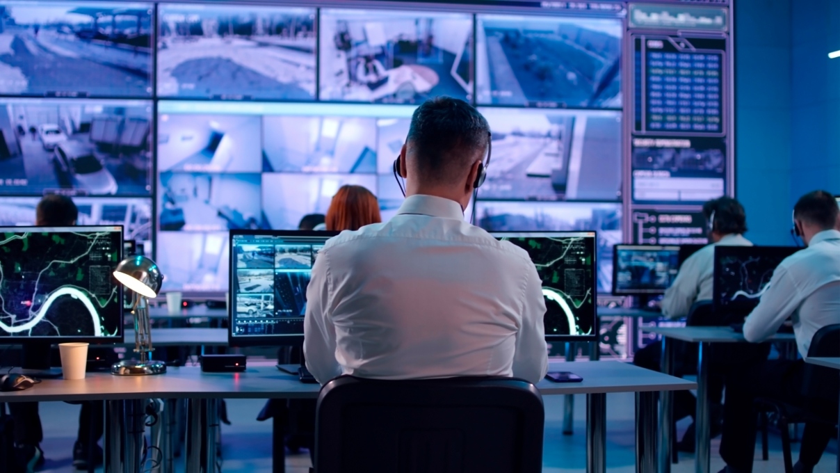 A physical security operator works at a central station monitoring multiple screens of video security footage.