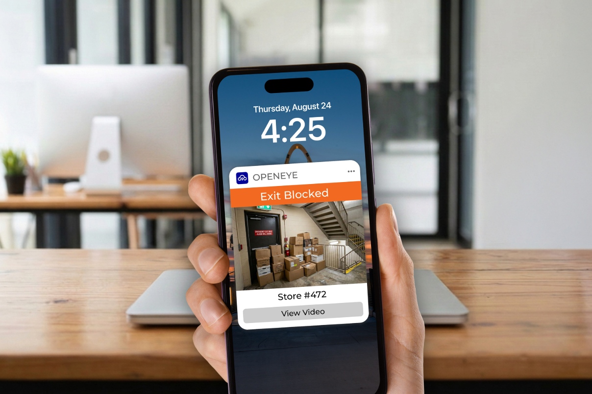A hand holds a phone that displays an OpenEye Web Services alert that reads "OPENEYE. Exit Blocked. Store #472. View Video."