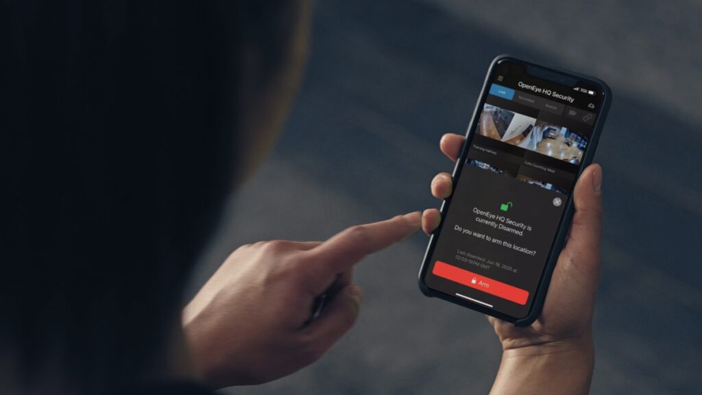 A person holds a mobile phone which displays OpenEye Web Services's Virtual Arm/Disarm feature.