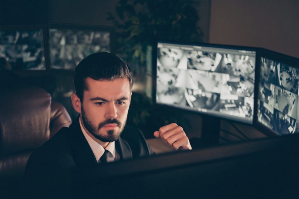 A man in a suit watches several computer monitors displaying video security footage.